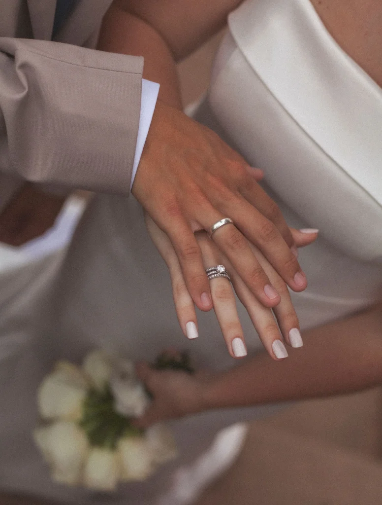 Close-up of the couple’s hands showing their wedding rings, with the bride holding a bouquet of white roses.