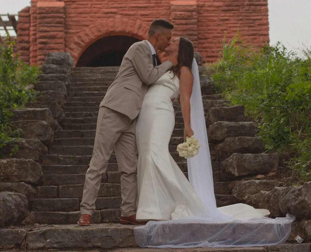 Bride and groom kissing on stone steps, with the groom dipping the bride as she holds a bouquet of white roses.