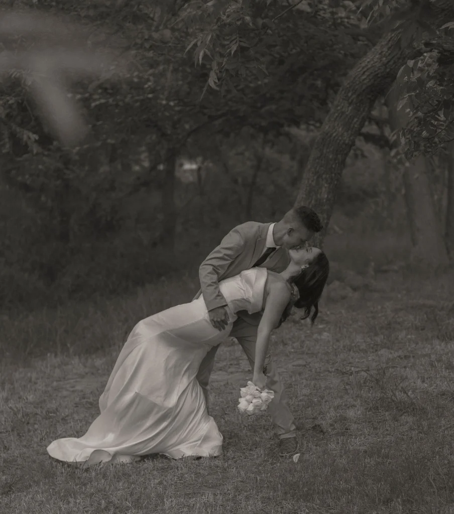 Black and white photo of groom dipping the bride backward for a kiss in a wooded outdoor setting.