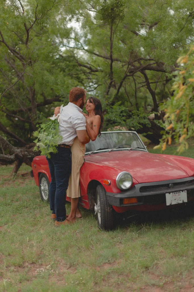 Man holding a bouquet and embracing a smiling woman as she leans against a vintage red convertible in a forest clearing.