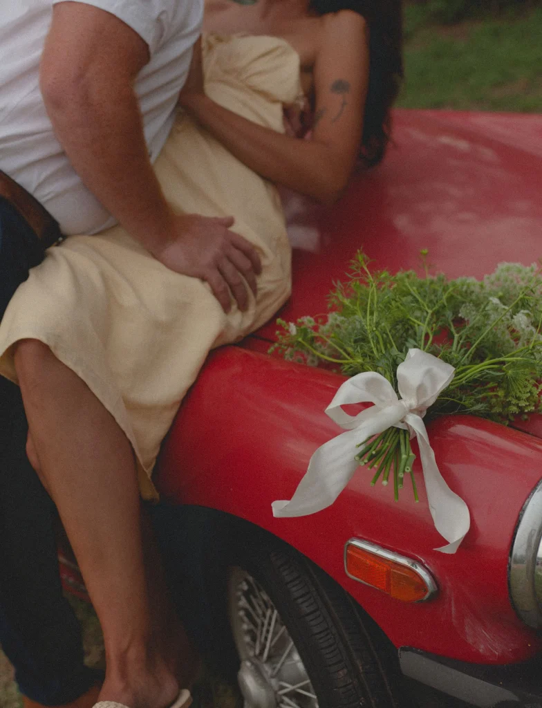 Close-up of the couple sitting on the red car hood with a bouquet tied with a white ribbon resting beside them.