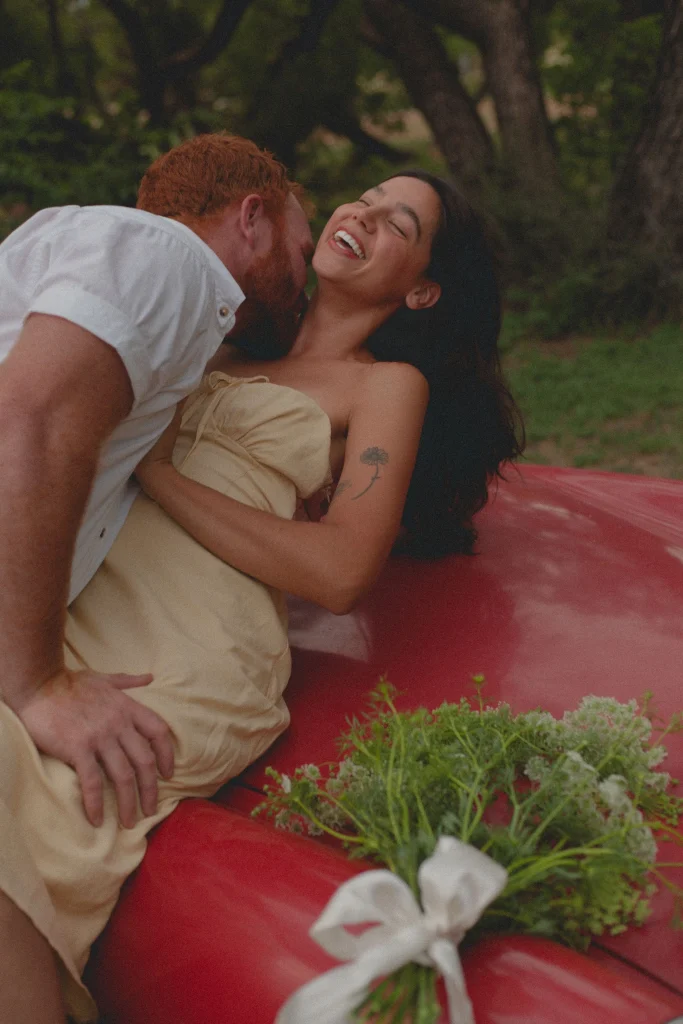 Man leaning over a laughing woman as they lie on the hood of a red convertible with a bouquet nearby.