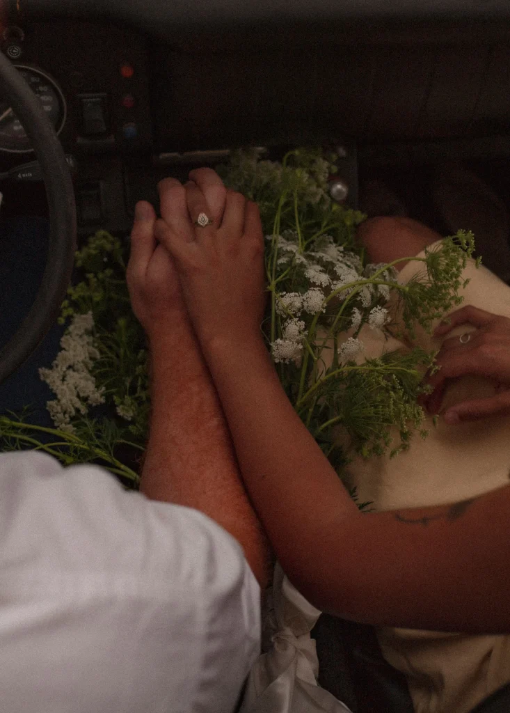 Close-up of the couple holding hands inside the car with flowers resting across their laps.