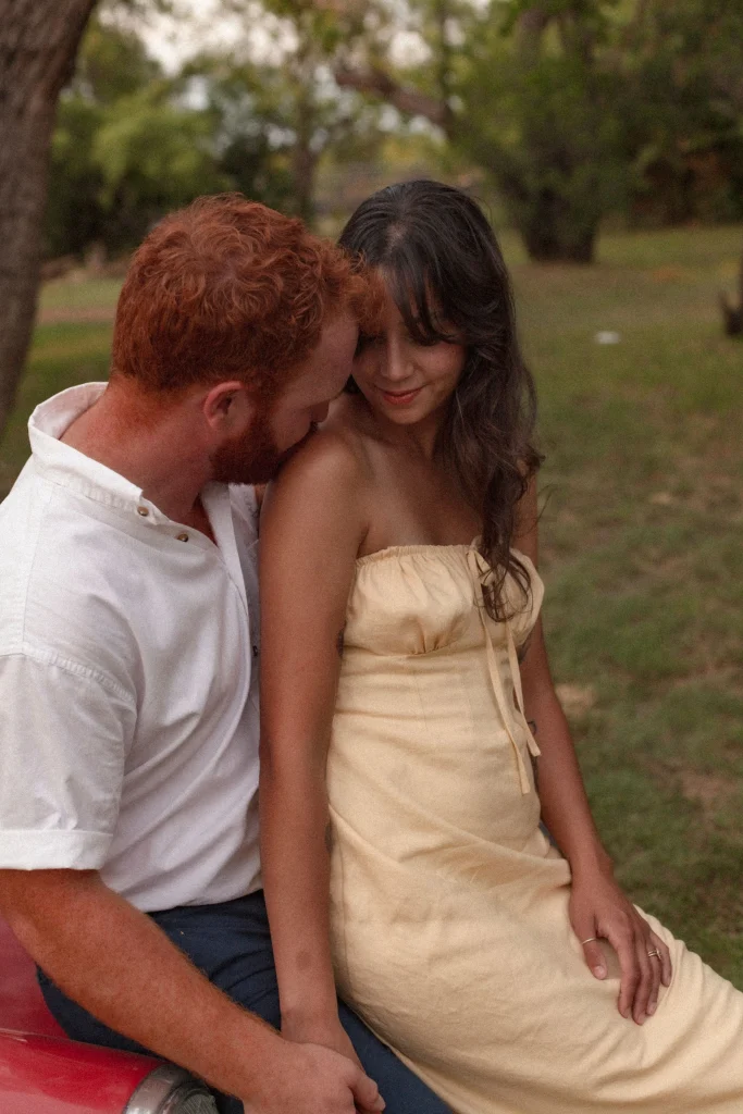 Man sitting close to a woman in a yellow dress as they share an intimate moment beside a red convertible.