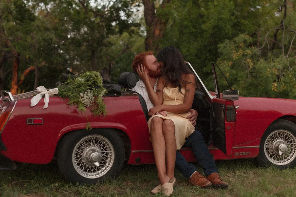 Couple kissing while sitting in a red vintage convertible decorated with flowers in a wooded area.