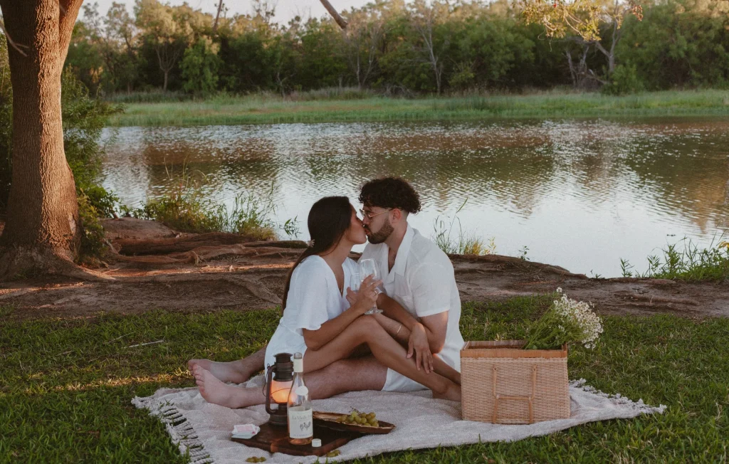 Couple kissing during a lakeside picnic with wine and flowers at sunset.