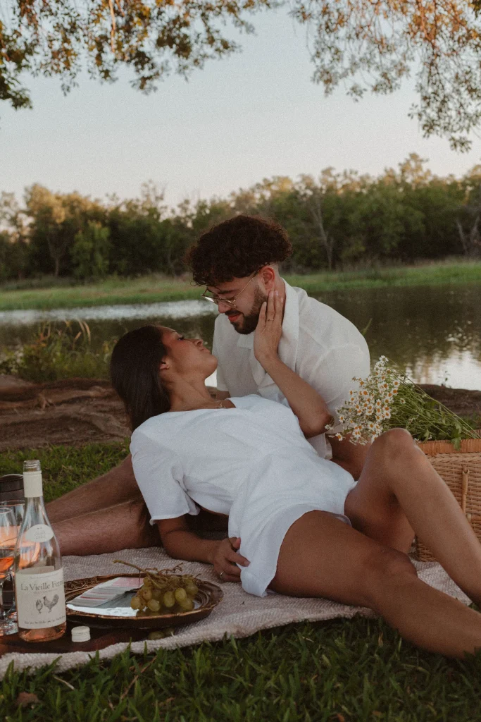 Couple lounging on a picnic blanket by a lake, looking into each other's eyes affectionately.