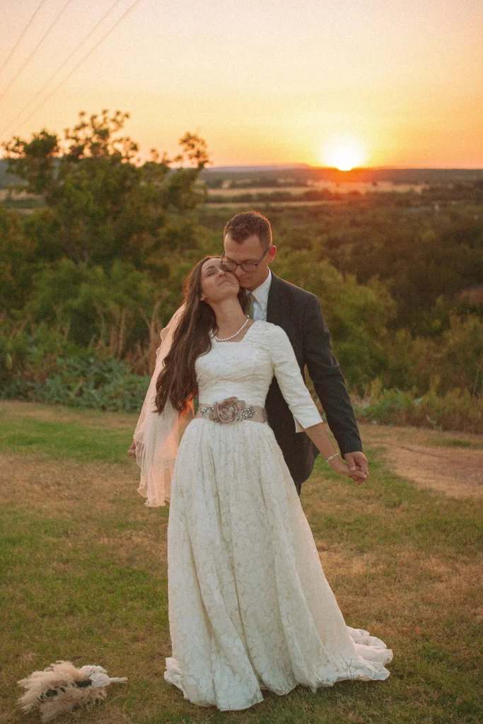 Groom kissing bride’s cheek as they stand in a field at sunset, holding hands in a romantic wedding moment.