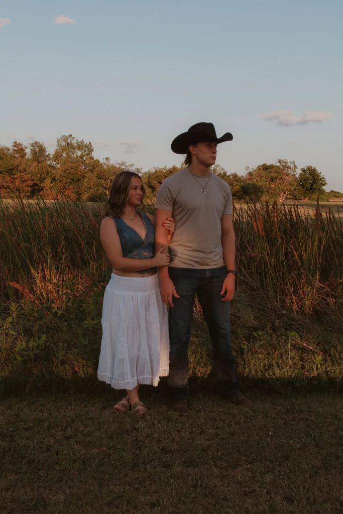 Couple standing together in a field at sunset, woman holding man’s arm while he wears a cowboy hat.