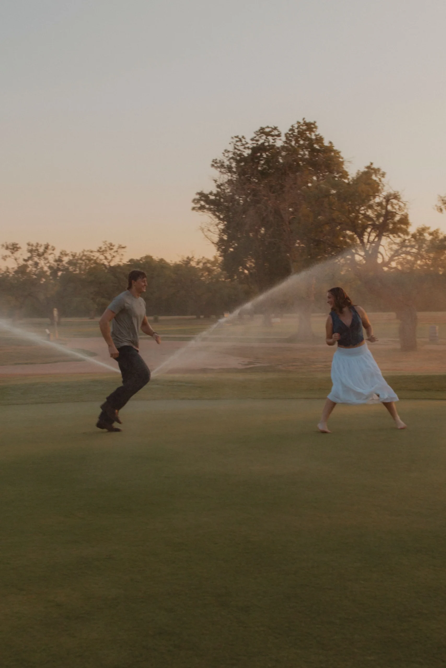 Couple running and laughing through sprinklers on a golf course at dusk.