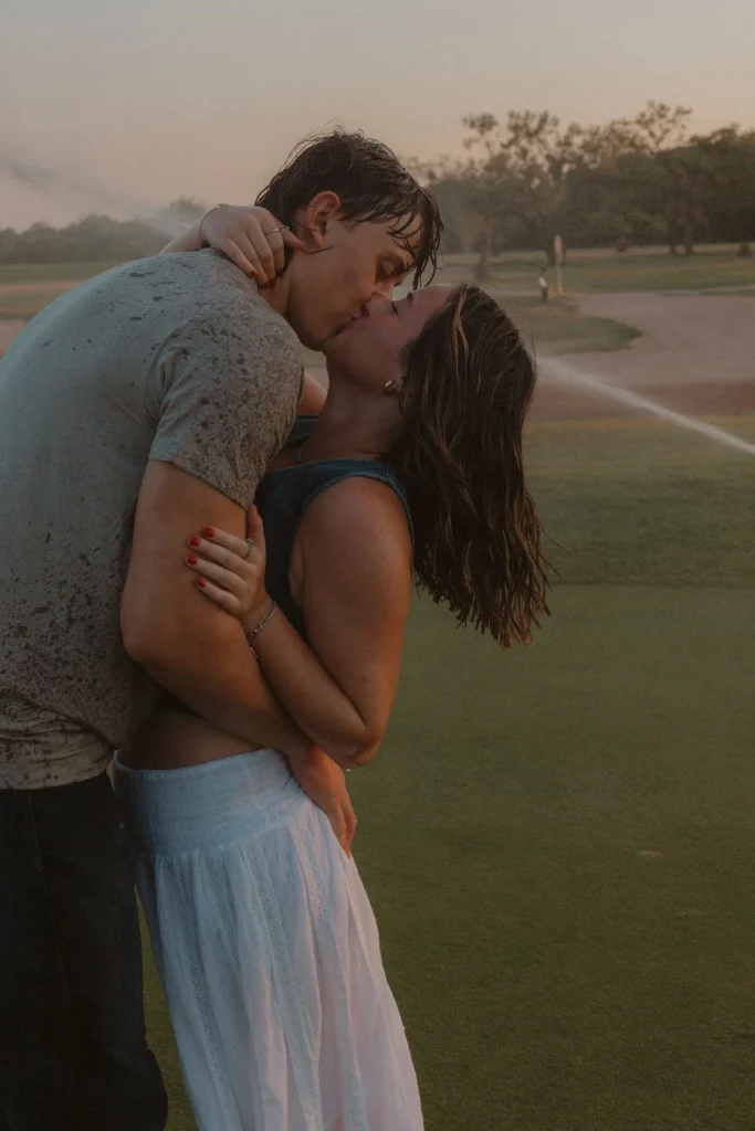 Wet couple sharing a kiss on a golf course with sprinklers spraying behind them.