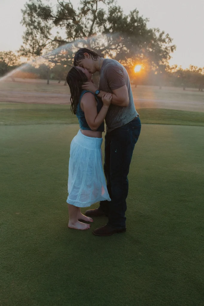 Couple embracing and kissing on a golf course at sunset while soaked from sprinklers.