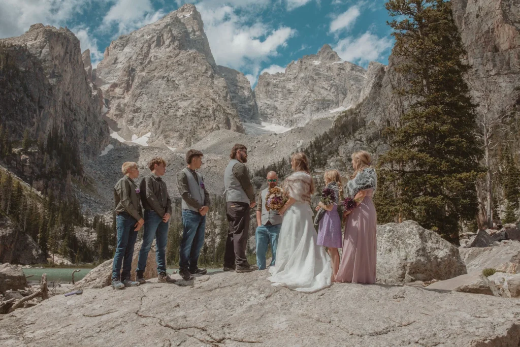Wedding ceremony taking place on a rocky mountain ledge overlooking a lake and towering peaks.