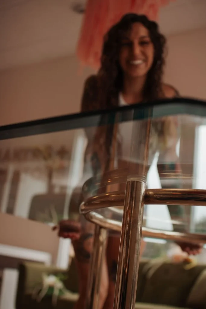 Close-up of a woman standing behind a glass table, wearing white underwear, showing tattoos on her thighs and arms.