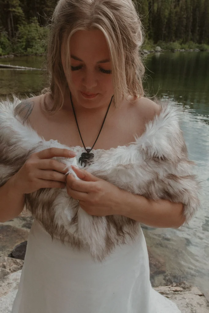 Bride adjusting her fur shawl while standing beside a calm alpine lake.