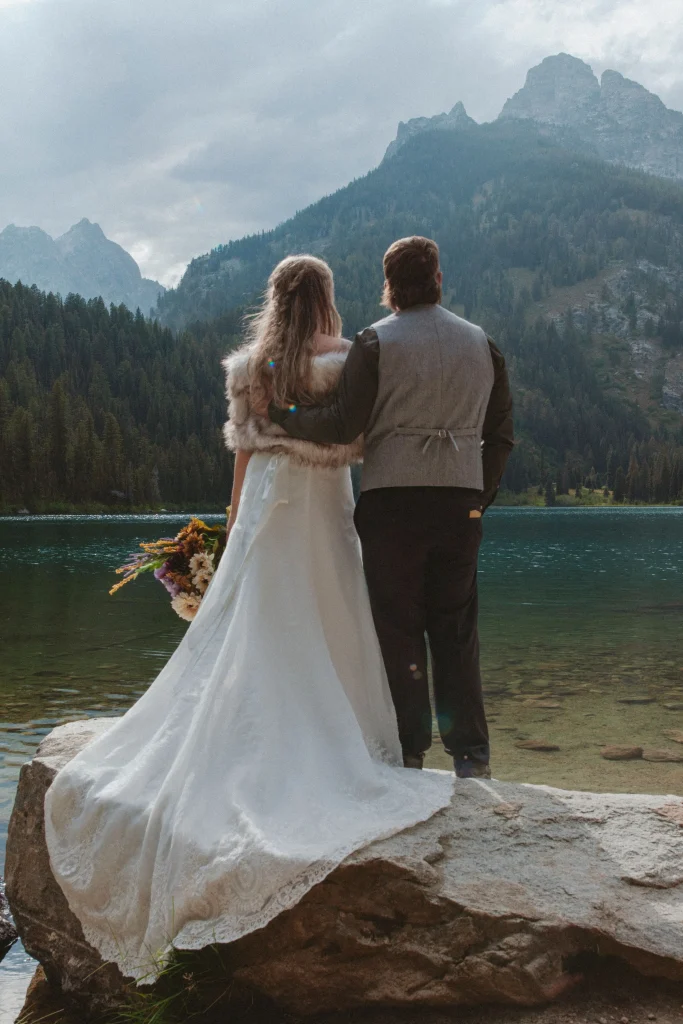 Bride and groom standing arm in arm on a rock overlooking a mountain lake.