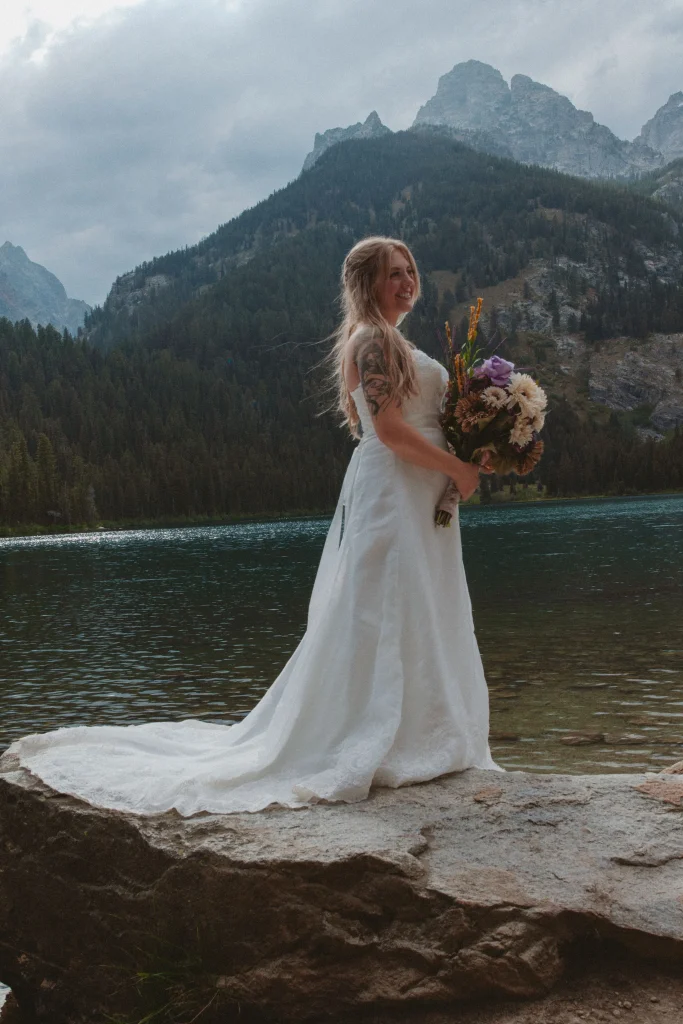 Bride standing alone on a rock with bouquet in hand, smiling with mountains rising behind her.