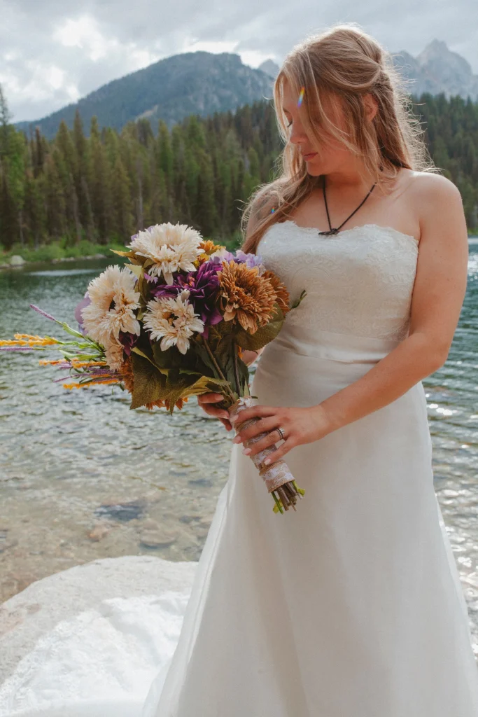 Bride holding a colorful bouquet while standing beside a mountain lake, looking down softly.