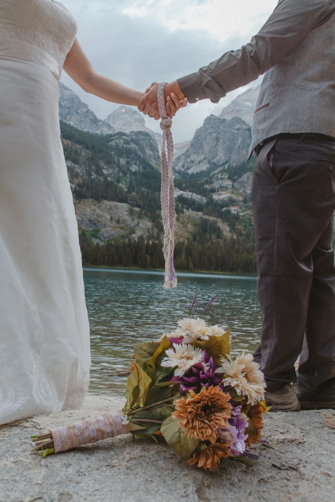 Bride and groom holding hands with a handfasting cord, bouquet resting on a rock by a mountain lake.
