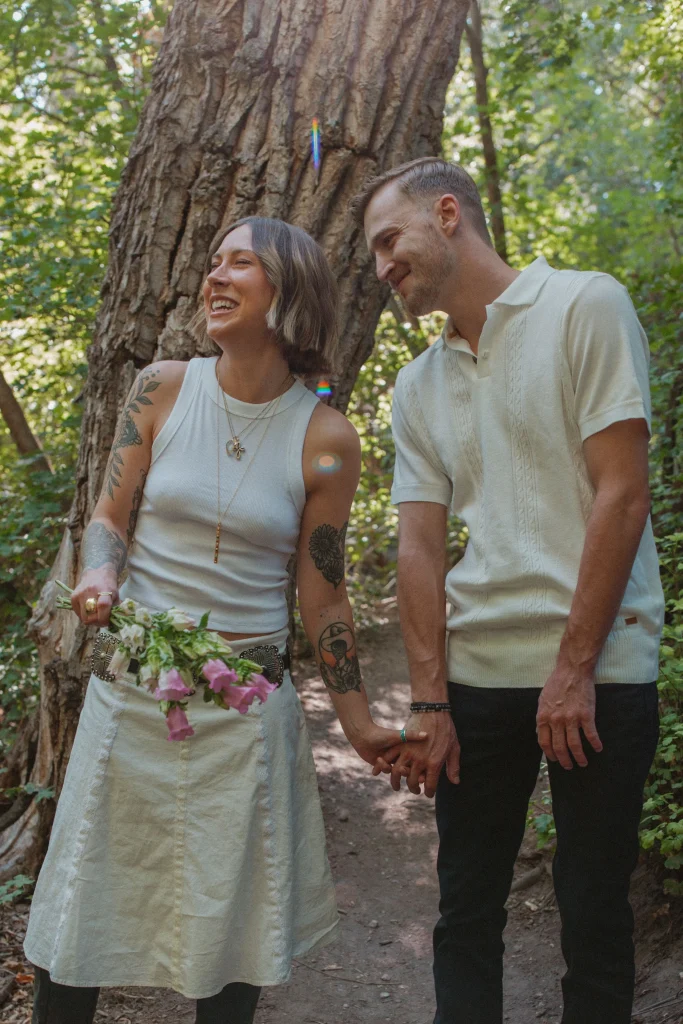 Couple holding hands and smiling together on a wooded trail, with woman holding flowers.