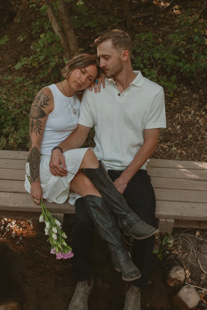 Couple sitting on a wooden bridge, with the woman resting her head on the man's shoulder while holding flowers.