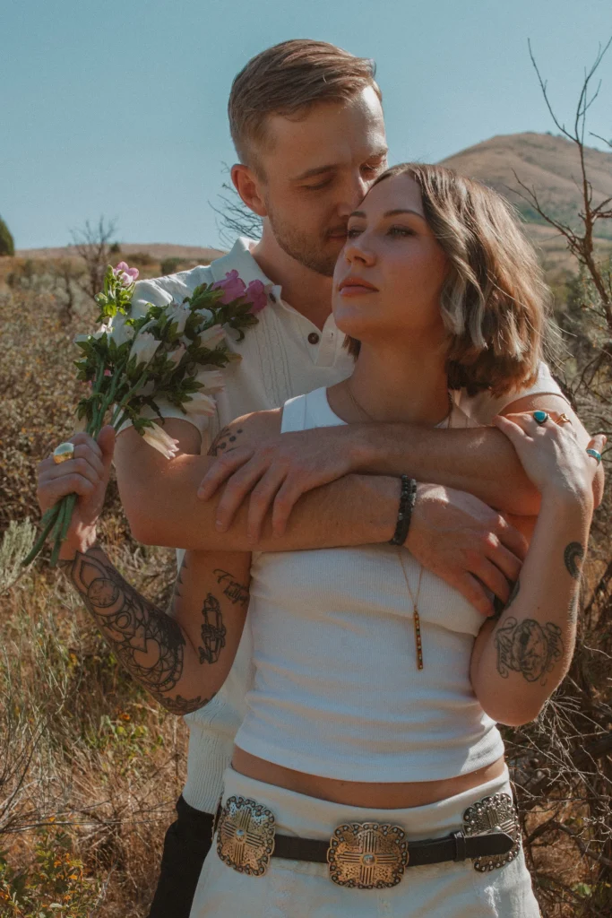 Man embracing a woman from behind in a sunlit field while she holds a bouquet of flowers.