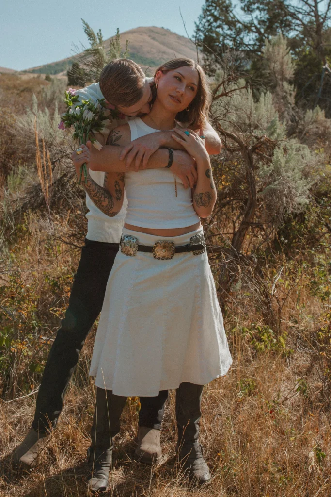 Couple standing in a golden field as the man kisses the woman’s neck while she holds flowers.