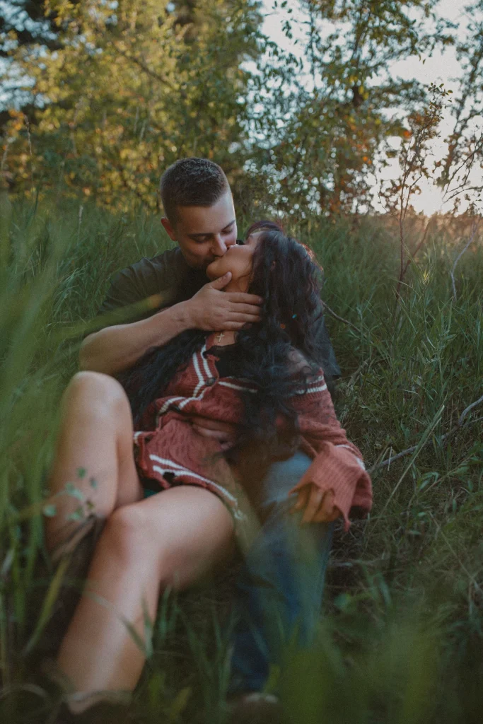 Couple kissing while sitting together in tall grass during golden hour.