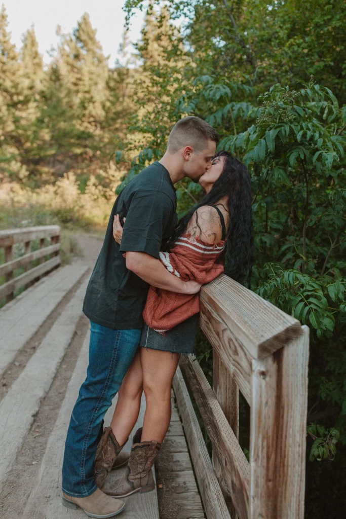 Couple kissing while leaning against a wooden bridge railing surrounded by trees.