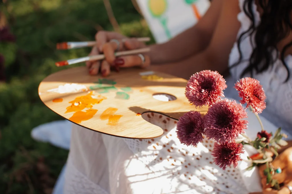 Close-up of an artist's paint palette with colorful paints beside purple flowers in sunlight.