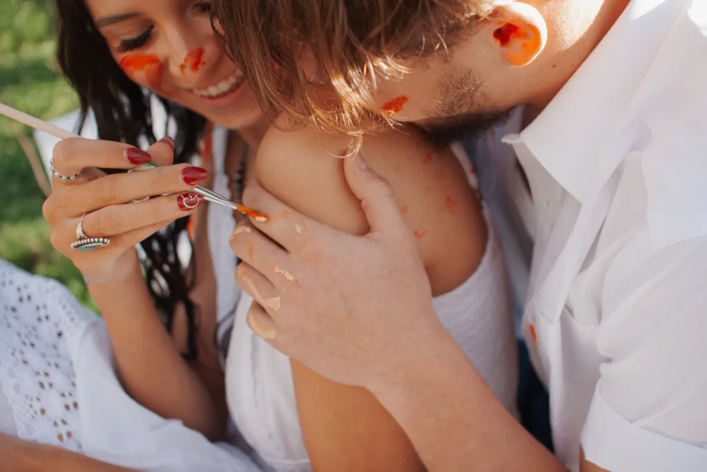 Couple laughing and painting on each other’s skin during a playful outdoor paint session.