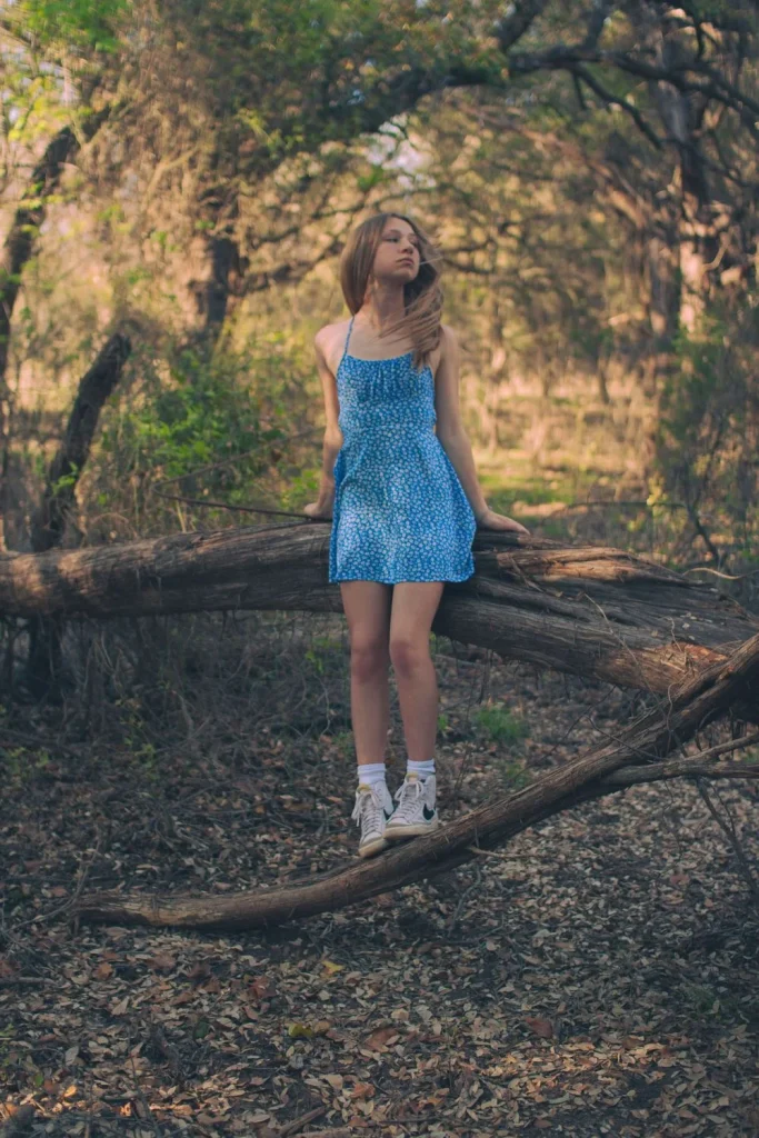 Girl in a blue floral dress standing on a fallen tree in a wooded area with sunlight filtering through the branches.
