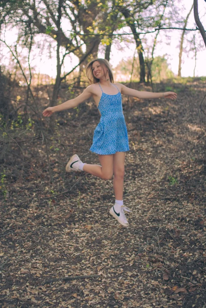 Girl in a blue dress joyfully twirling on a forest path covered in leaves.