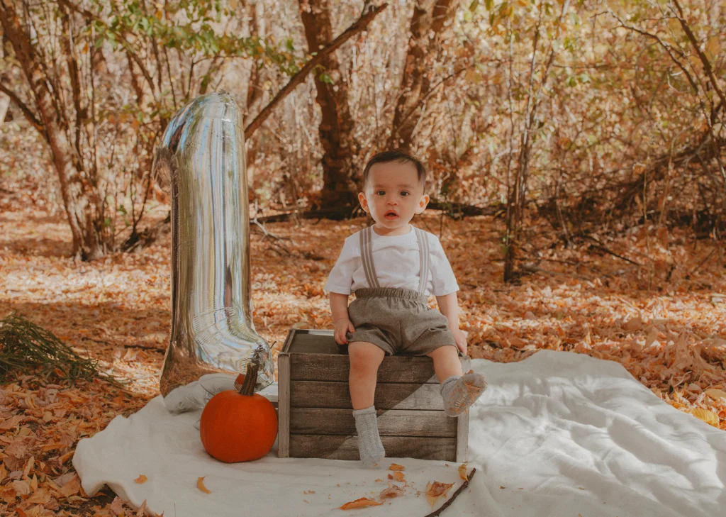Baby sitting on a wooden crate outdoors with a pumpkin and a silver number one balloon.