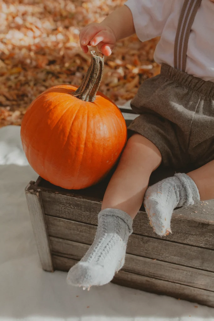 Close-up of a baby’s hand touching a pumpkin next to their feet.
