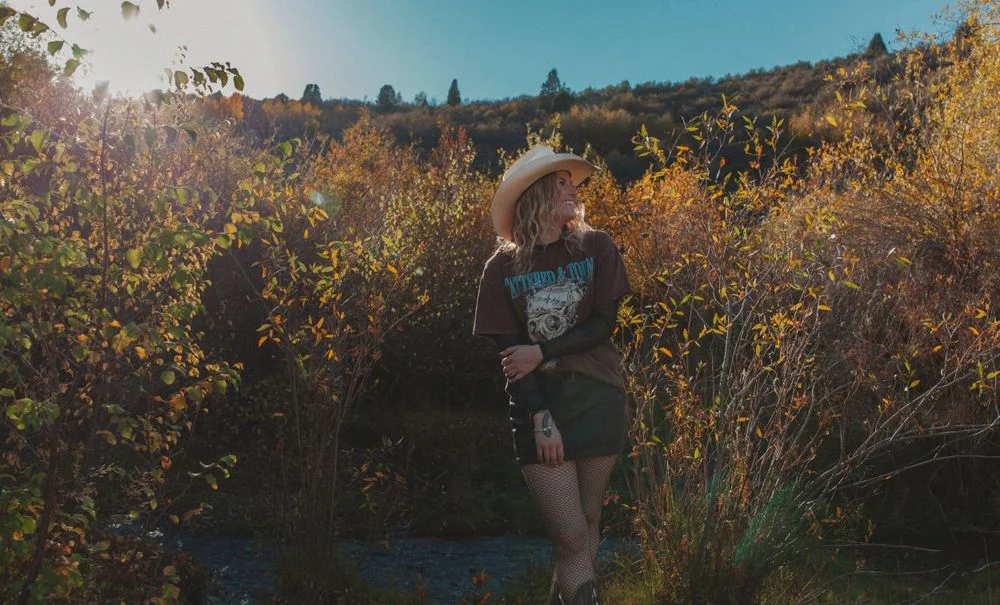 Woman in a cowboy hat standing among autumn-colored bushes with bright sunlight behind her.