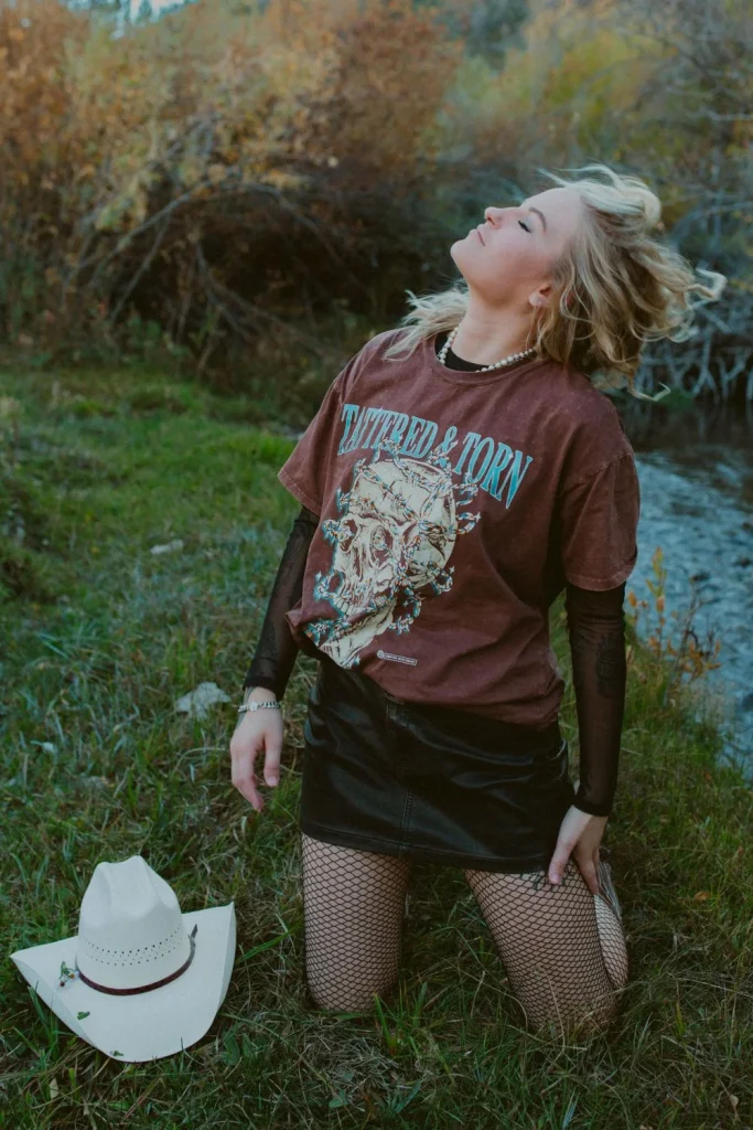 Woman kneeling in the grass beside a white cowboy hat, head tilted back, wearing a graphic tee and fishnet tights.