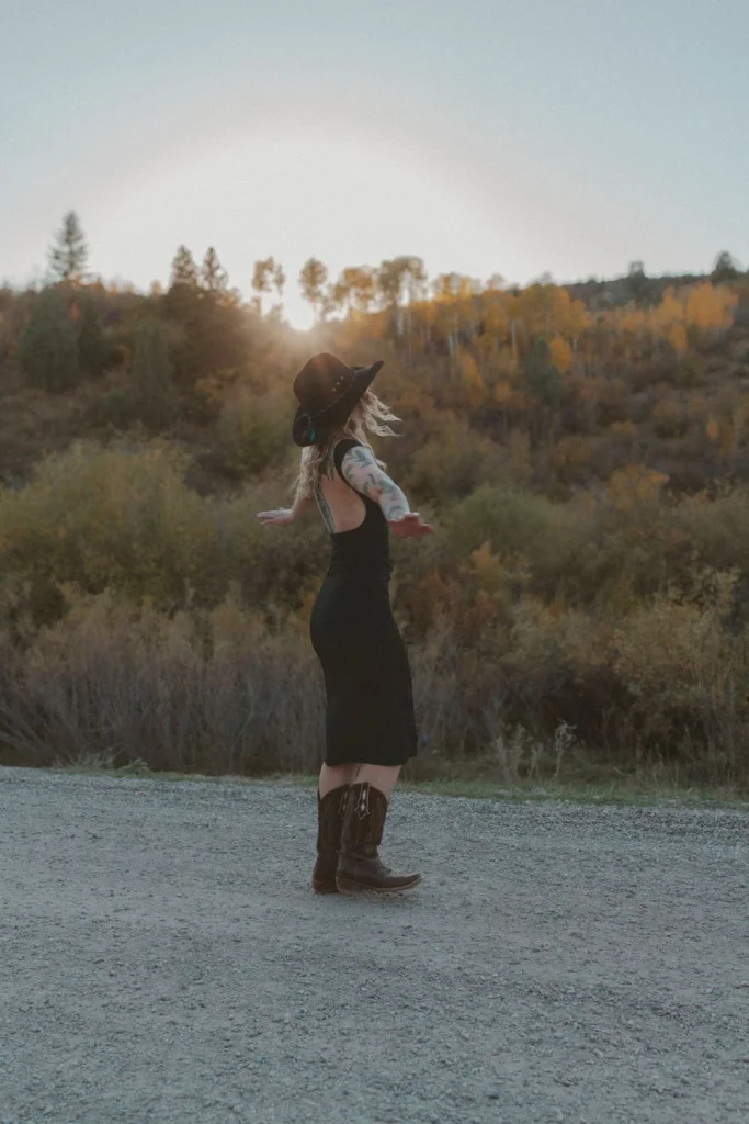 Woman in a black dress and cowboy boots spinning on a gravel road with sunset light behind her.