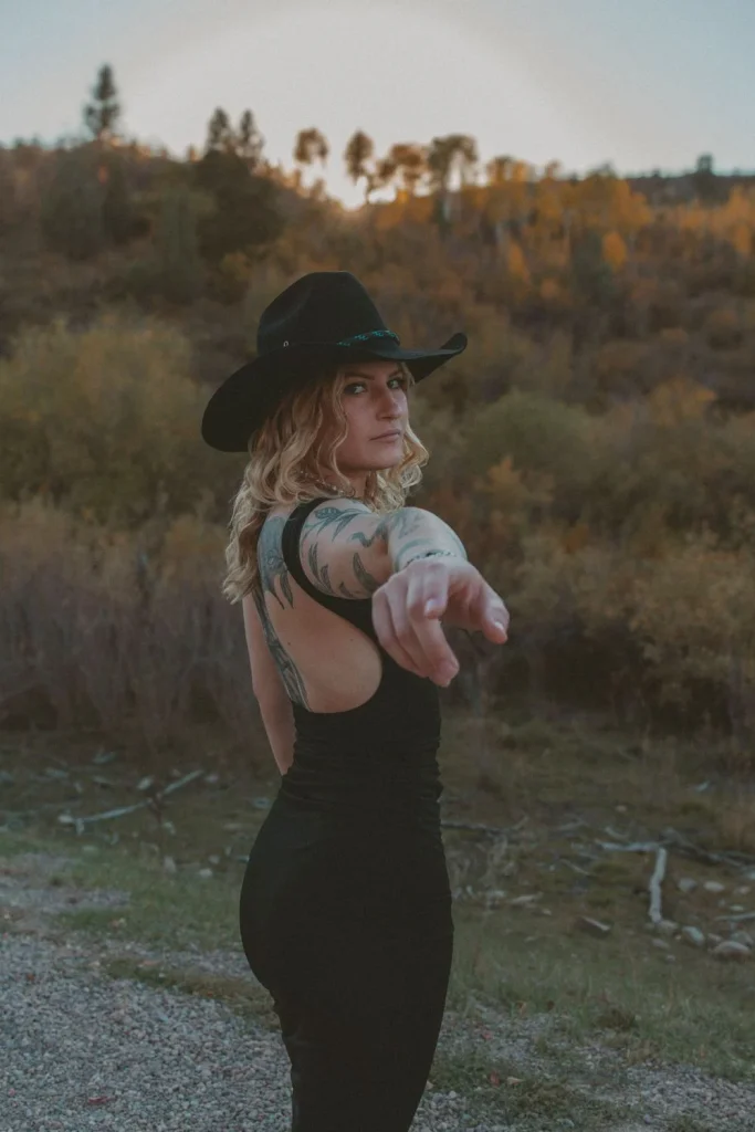 Woman in a black dress and cowboy hat pointing toward the camera with autumn hills in the background.