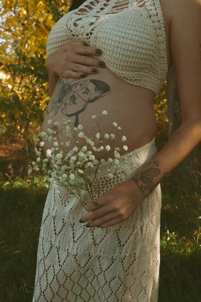 Pregnant woman holding her belly and a bouquet of baby’s breath flowers, showing tattoos.