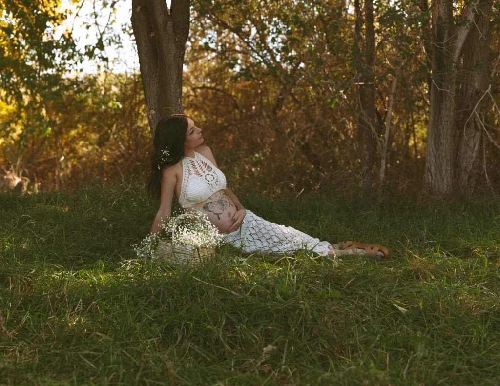 Pregnant woman sitting under a tree in a crocheted outfit with a basket of baby’s breath.