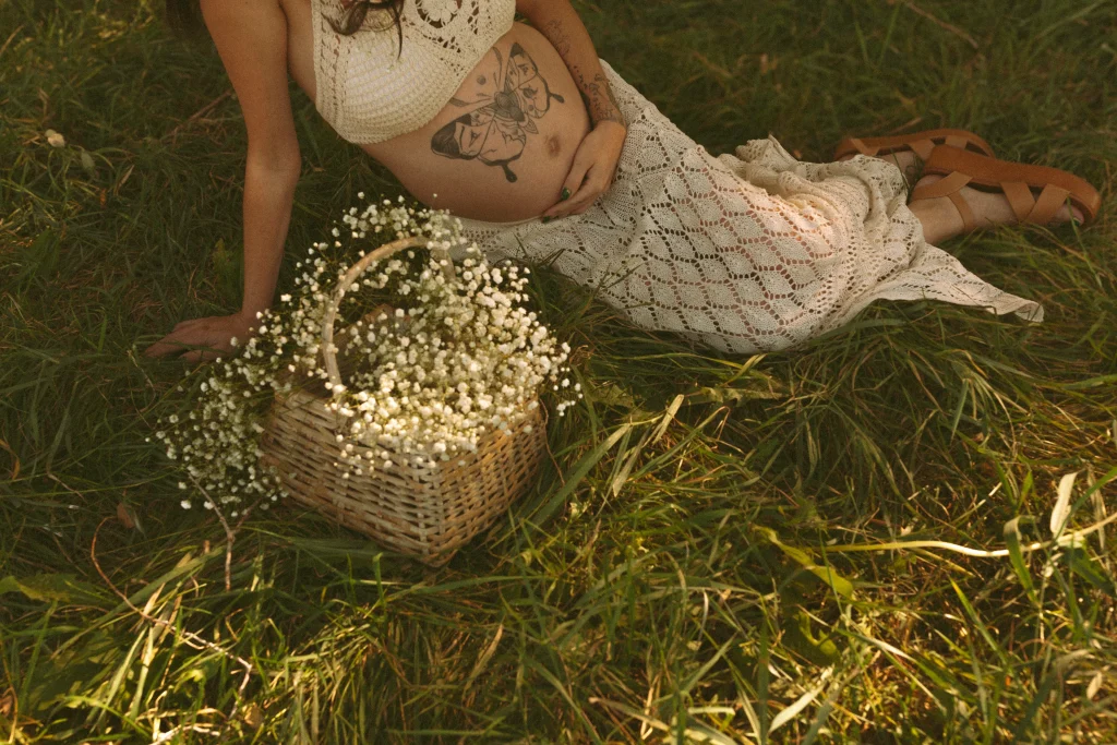 Basket of baby’s breath flowers resting in grass beside a pregnant woman.