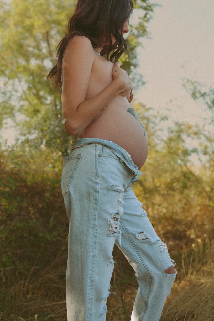 Pregnant woman standing topless in ripped jeans, cradling her belly outdoors.