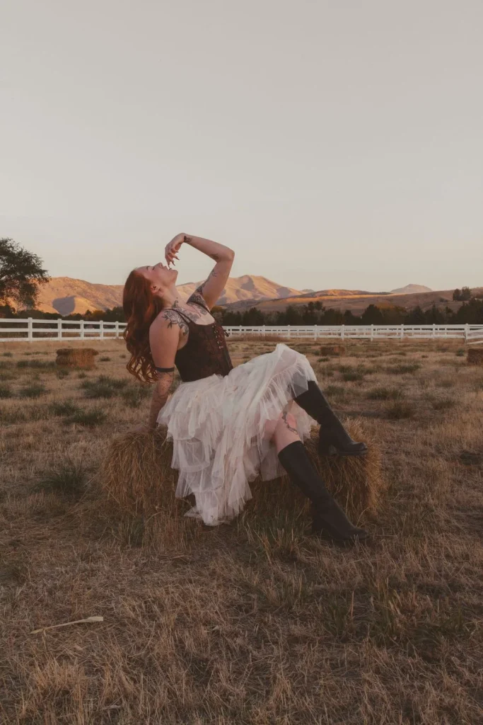 Woman sitting on a hay bale in a dry field at golden hour, wearing a corset and layered tulle skirt, posing with her head tilted back.