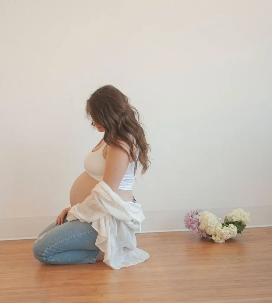Pregnant woman kneeling on a wooden floor, looking down at her belly with flowers beside her.