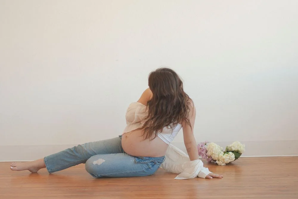 Pregnant woman reclining on the floor in jeans and a white top, with flowers arranged behind her.