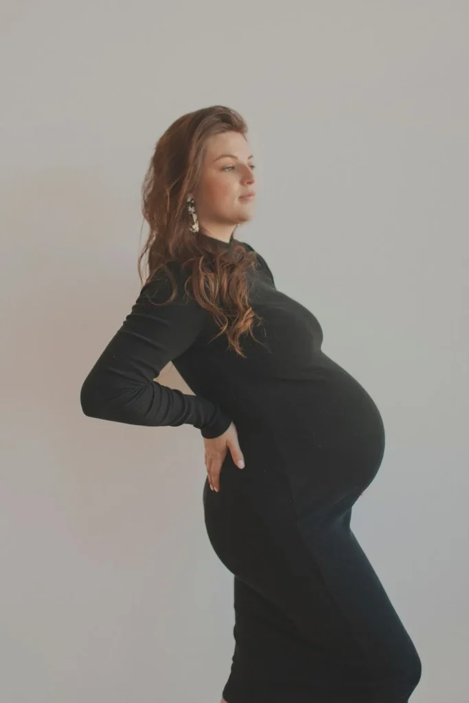 Pregnant woman in a fitted black dress standing sideways with one hand on her hip.