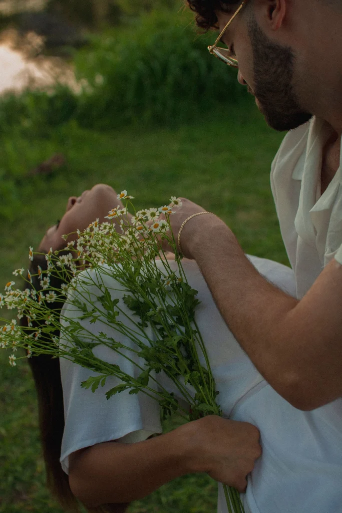Man holding a woman dipped back while she holds a bouquet of daisies in a grassy field.