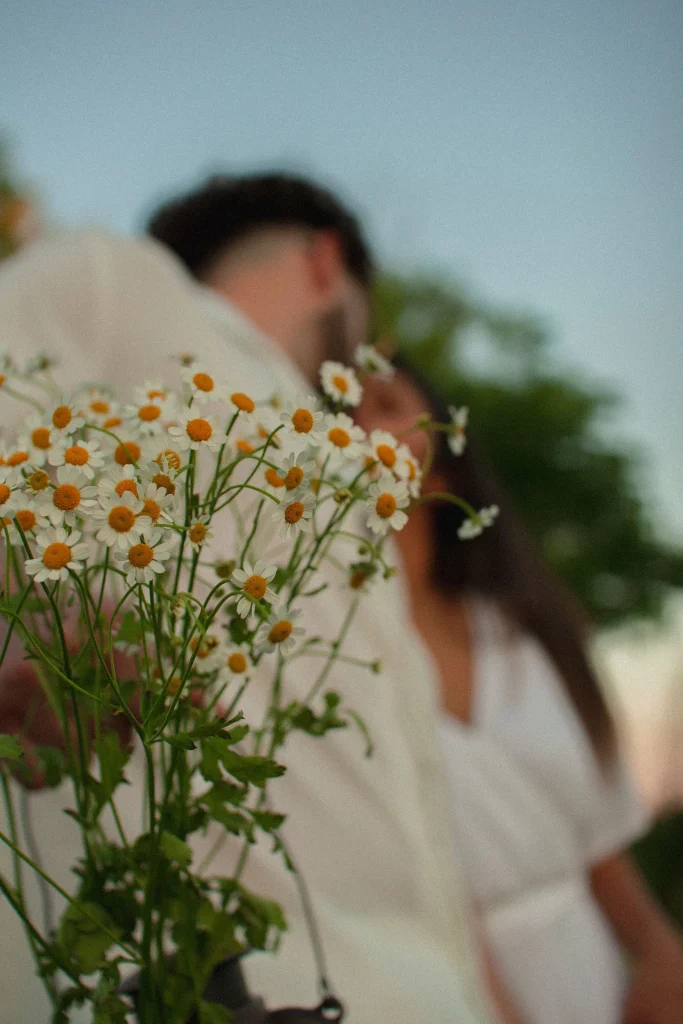 Close-up of daisies in the foreground as a couple leans in toward each other wearing white outfits.