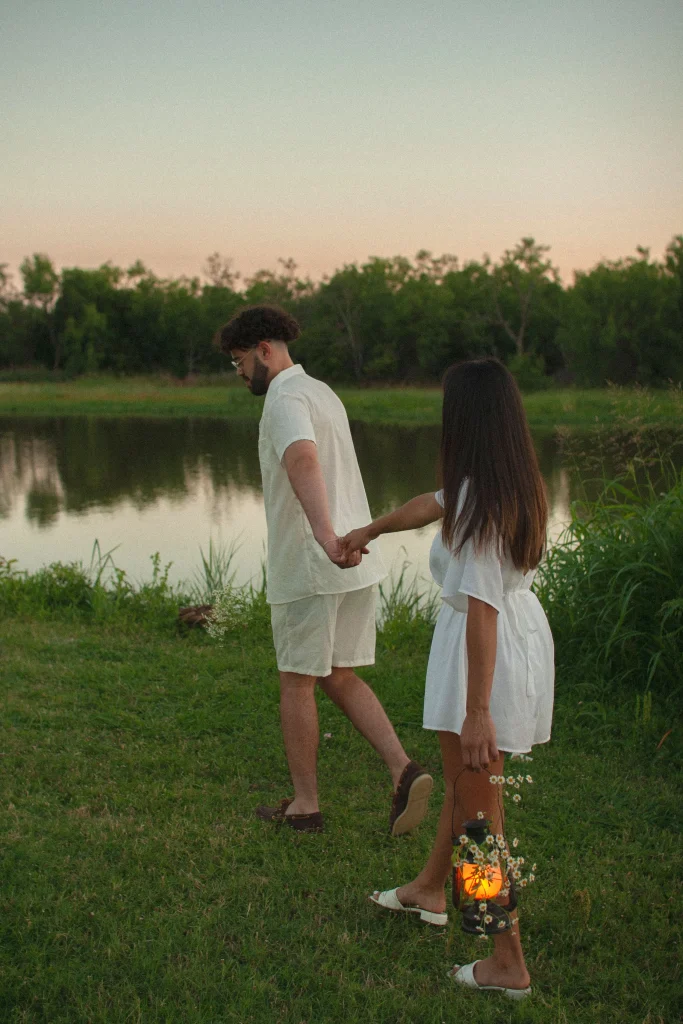 Couple walking hand in hand along a lakeside at sunset, with the woman carrying a lantern decorated with daisies.